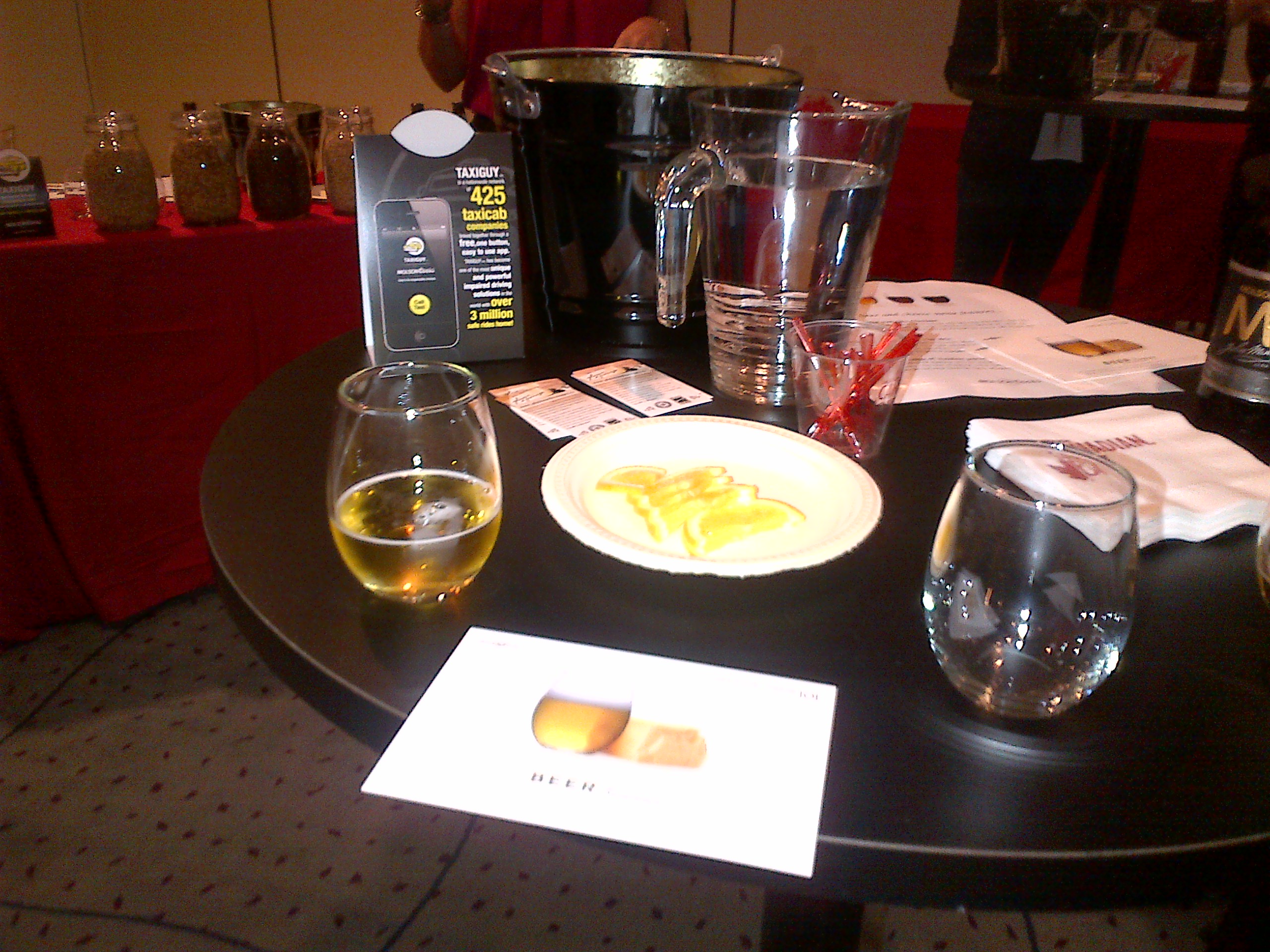 A hightop bar table with 1 small glass of beer, an empty glass, and a plate of cheese between the two glasses.