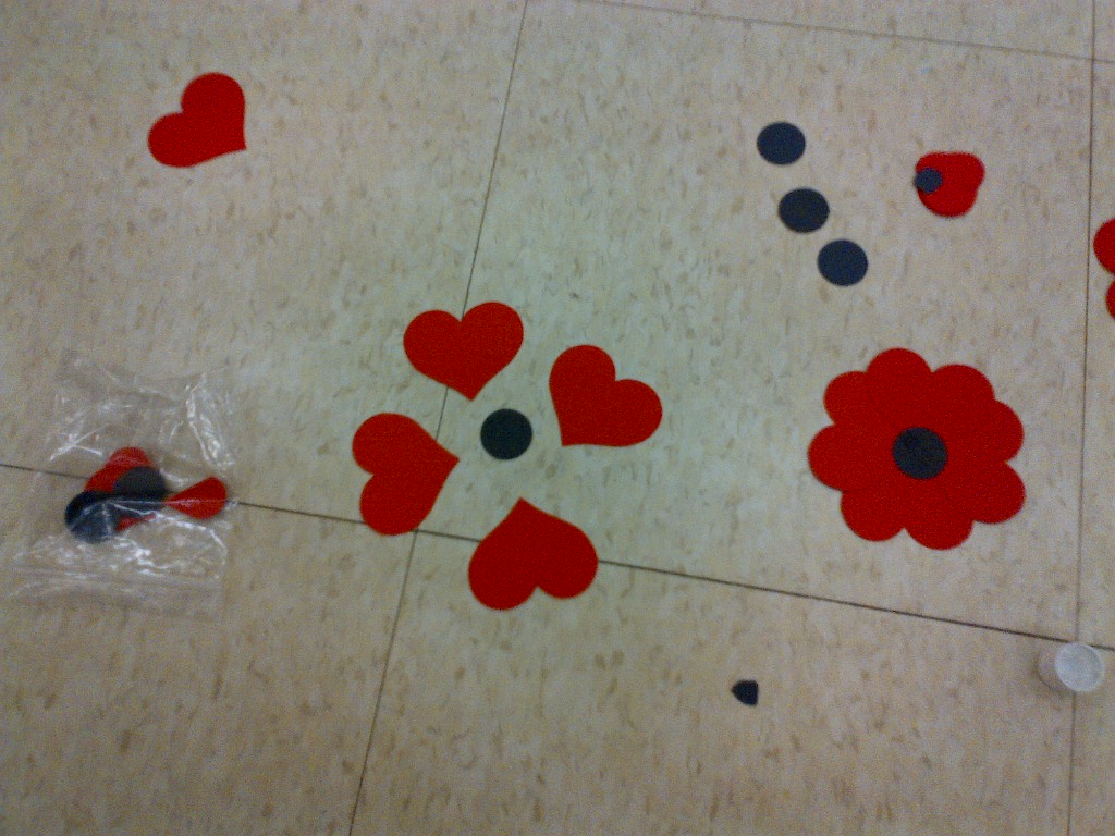 a view of 2 hand made poppies, one put together and one with the 4 hearts spaced about 4 cm apart from each other and the black circle in the middle. There are extra pieces scattered on the floor around the 2 poppies. 