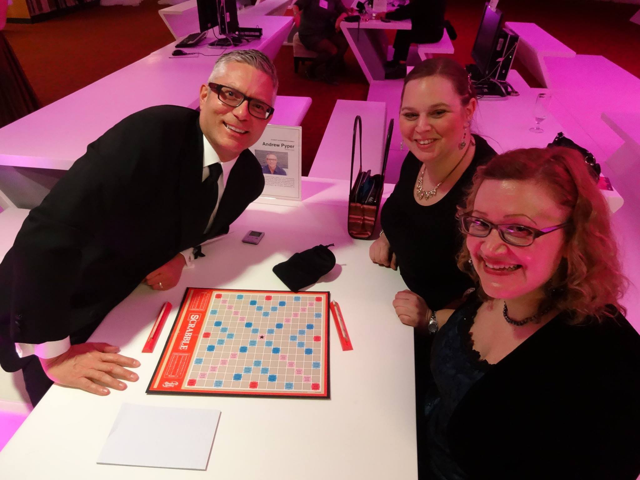 Two women sit opposite a middle aged man. An empty scrabble board is on the table between them