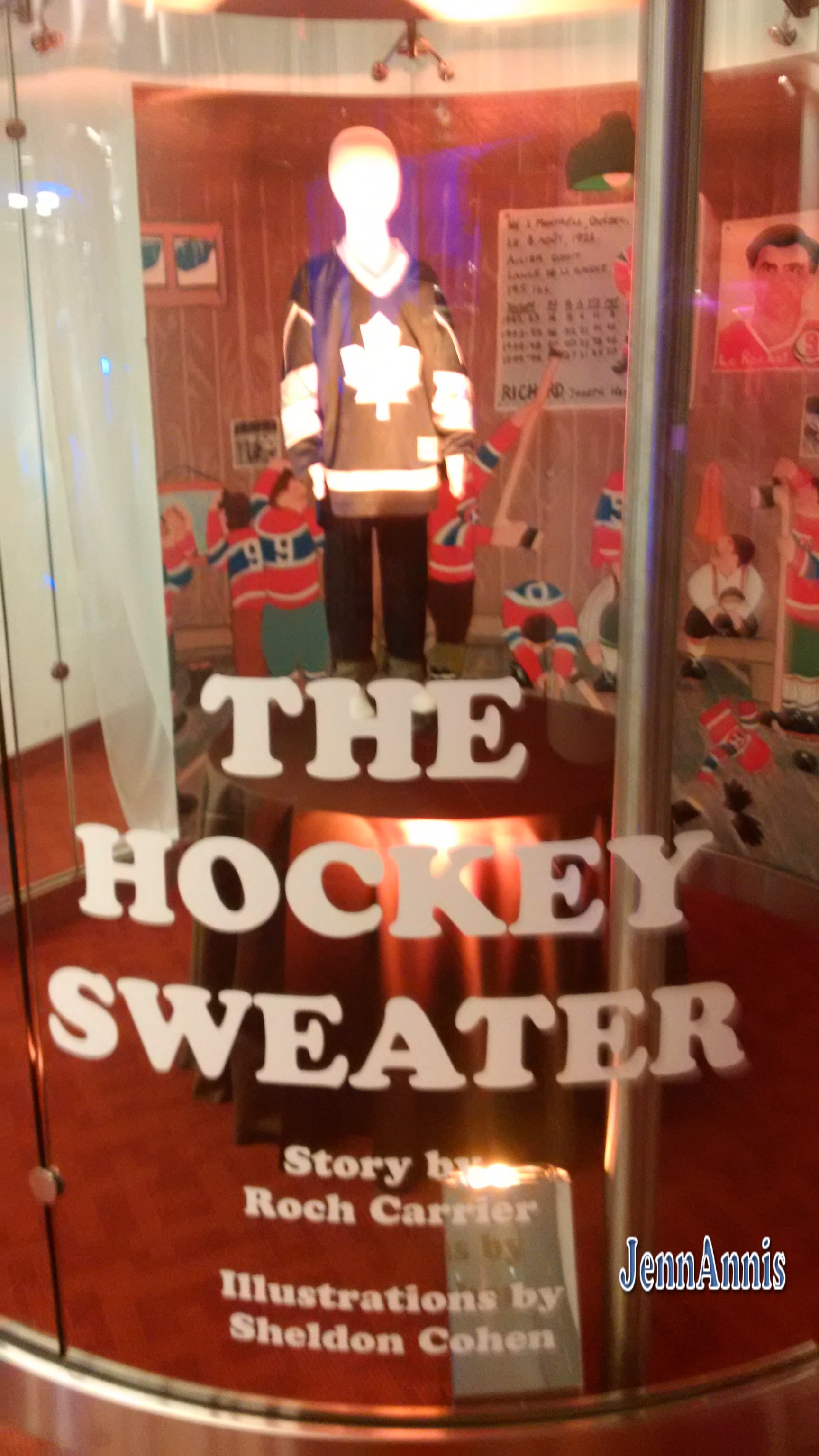 A mannequin dressed in a Toronto Maple Leafs jersey stands in front of a drawing of children all dressed in Montreal Canadians gear. Text reading: The Hockey Sweater