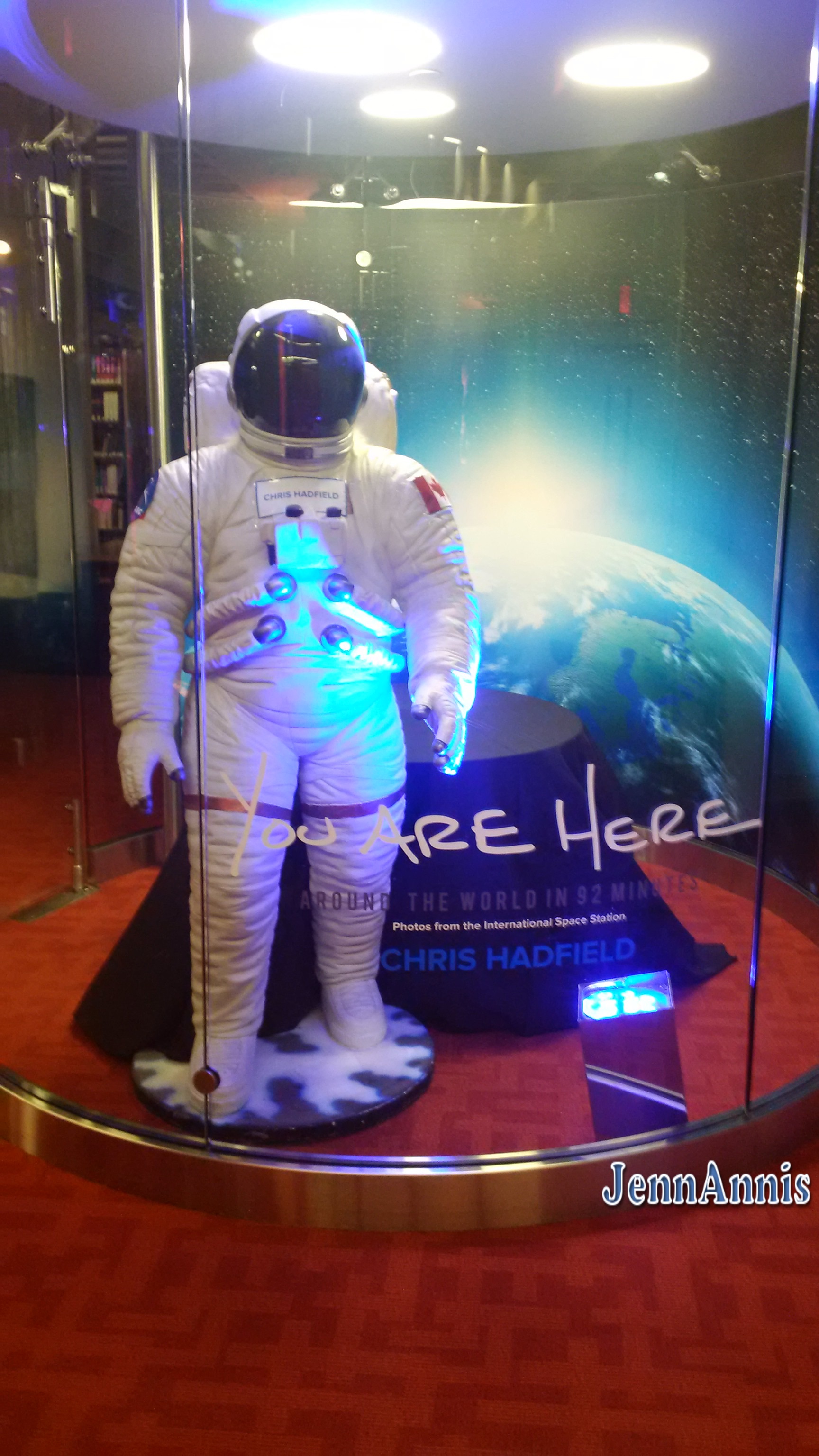 a Mannequin dressed in a white space suit and helmet stands in front of a cardboard earth. Text reading: You are here