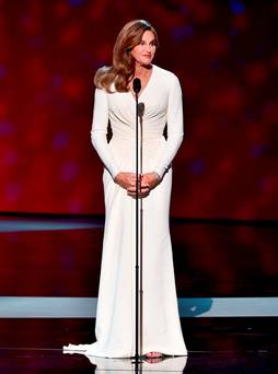Caitlyn Jenner at the mic at the ESPY awards. She is wearing a long white dress and smiling