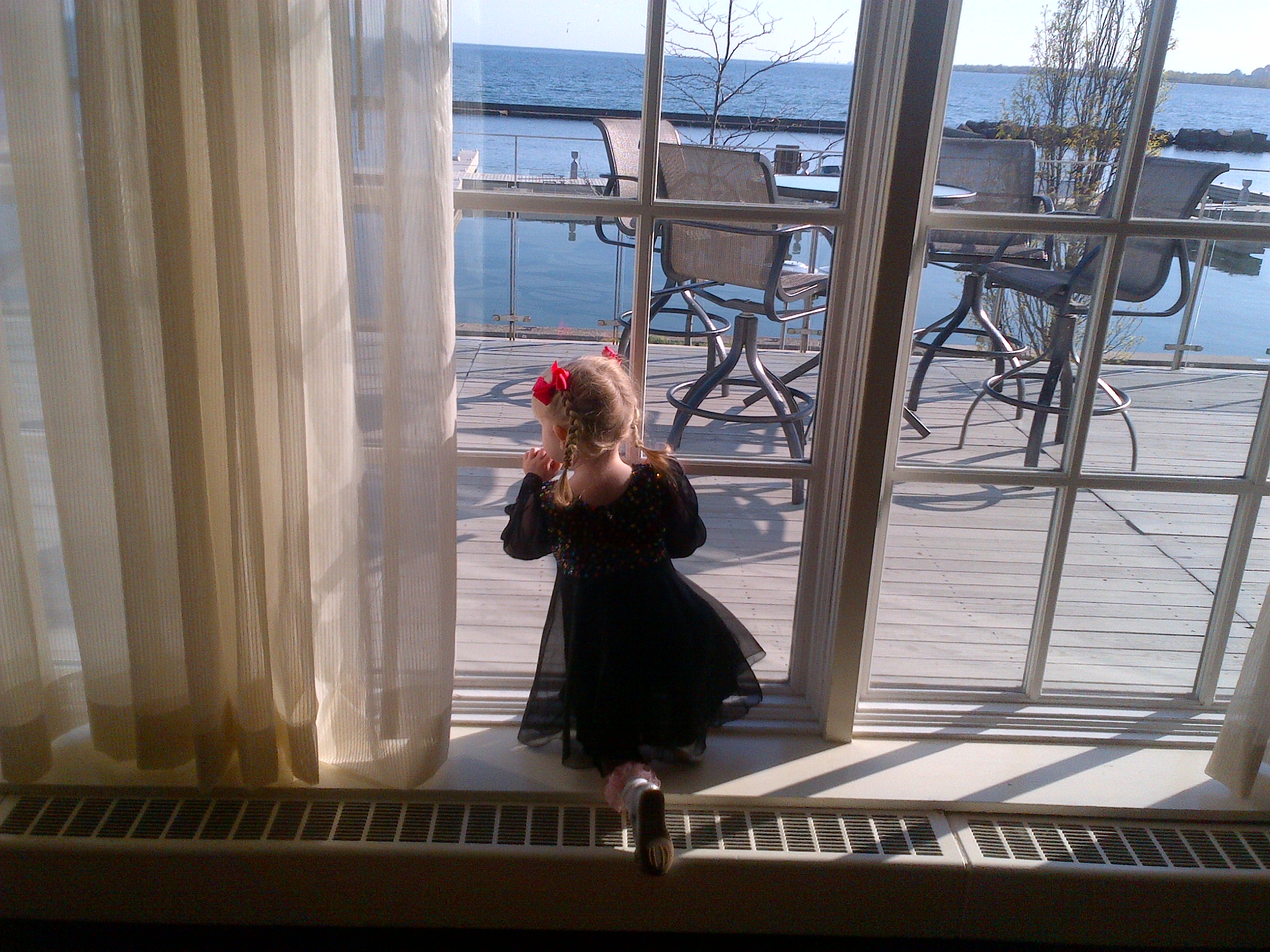 A 2 year old girl with blonde hair in pigtails with red bows at the top and wearing a fancy black dress looks out a window